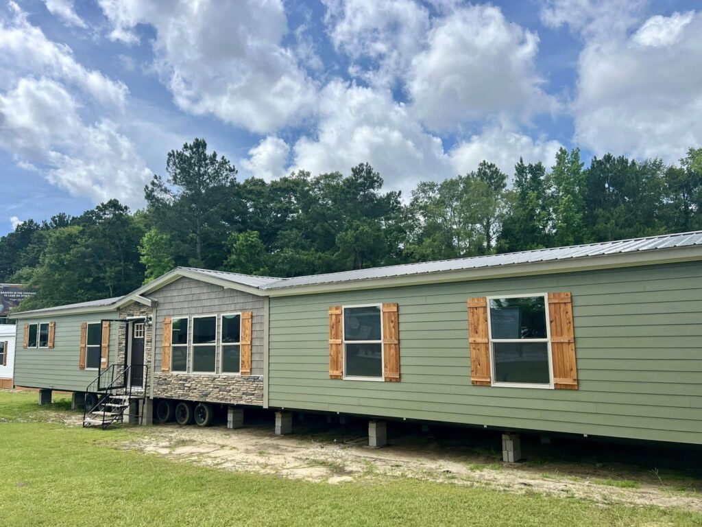 A large green manufactured home with wooden shutters and a stone accent is set among trees under a partly cloudy sky, conveying a serene, rustic vibe.