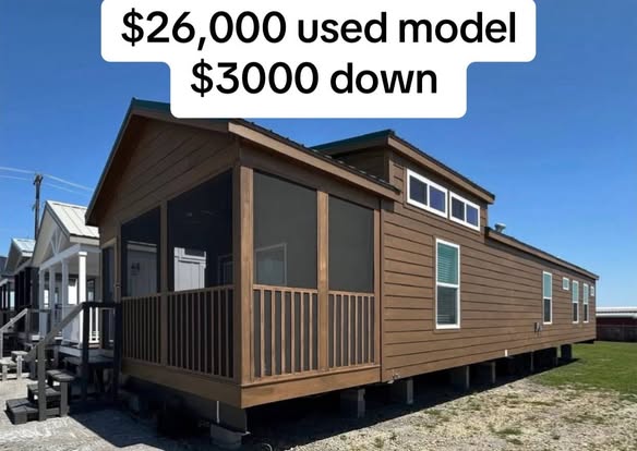 A brown mobile home with white-trimmed windows and a screened porch sits on a lot. Text above reads "$26,000 used model, $3000 down." Clear, sunny day.