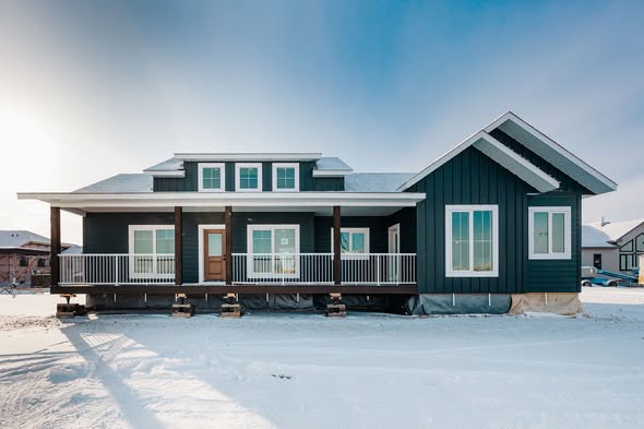 Blue house with white trim sits on a snowy landscape under a clear sky. It features a porch with railings and multiple windows, evoking a serene, wintery feel.
