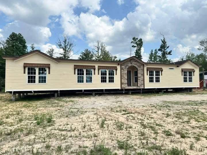 A beige manufactured home with large windows and a stone entrance sits on a sandy lot. Surrounded by trees under a partly cloudy sky, the scene is calm.