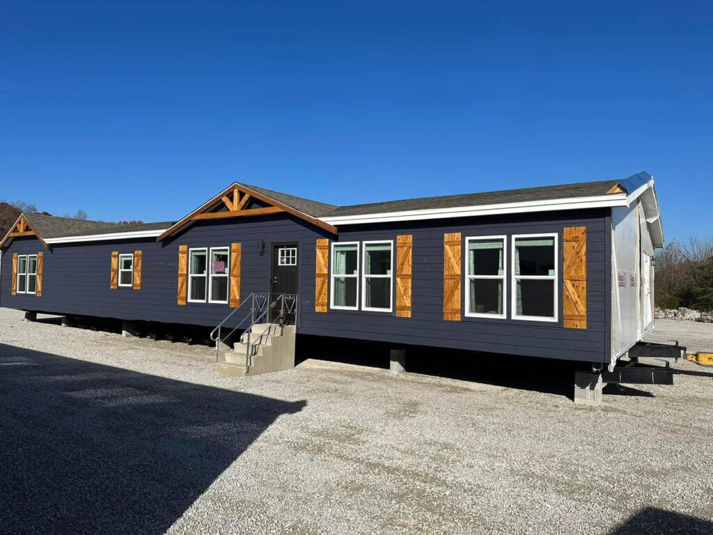 A modern, modular home with a dark gray exterior, wooden accents, and large windows sits on gravel. The clear blue sky adds a serene backdrop.