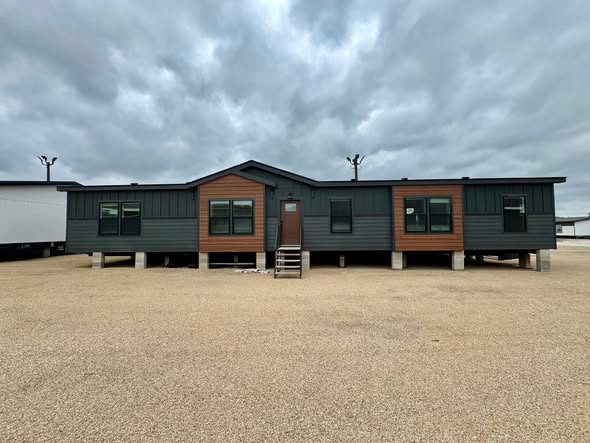 A modern, dark green modular home with wood accents sits elevated on a gravel lot. The structure is under a cloudy sky, conveying a subdued tone.