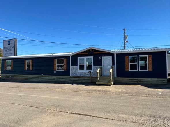 A blue manufactured home with white trim and a gabled roof against a clear sky. It has a small porch and a sign reading "Regional Homes" nearby.
