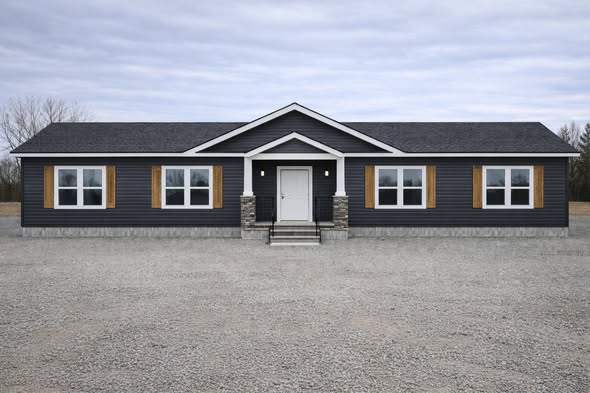 A modern, single-story house with dark blue siding, white trim, and symmetrical windows. It sits on a gravel lot under an overcast sky, creating a tranquil atmosphere.