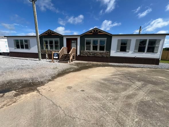 Single-story modular home with white siding and stone accents under a blue sky. Wooden steps lead to the door, surrounded by gravel and pavement.