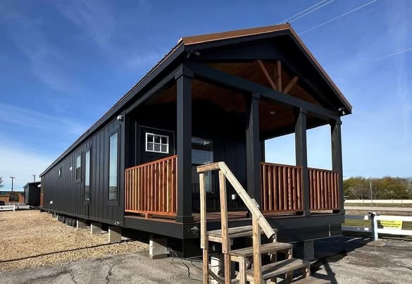 Modern black tiny house on stilts with a small wooden porch and steps, set against a clear blue sky. The design is sleek and minimalist.